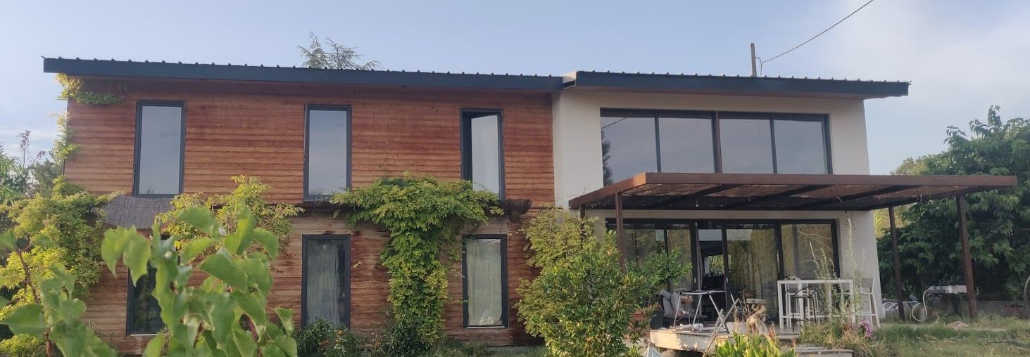 Maison bioclimatique autoconstruite : bardage bois, grandes baies vitrées et pergola, entourée de verdure